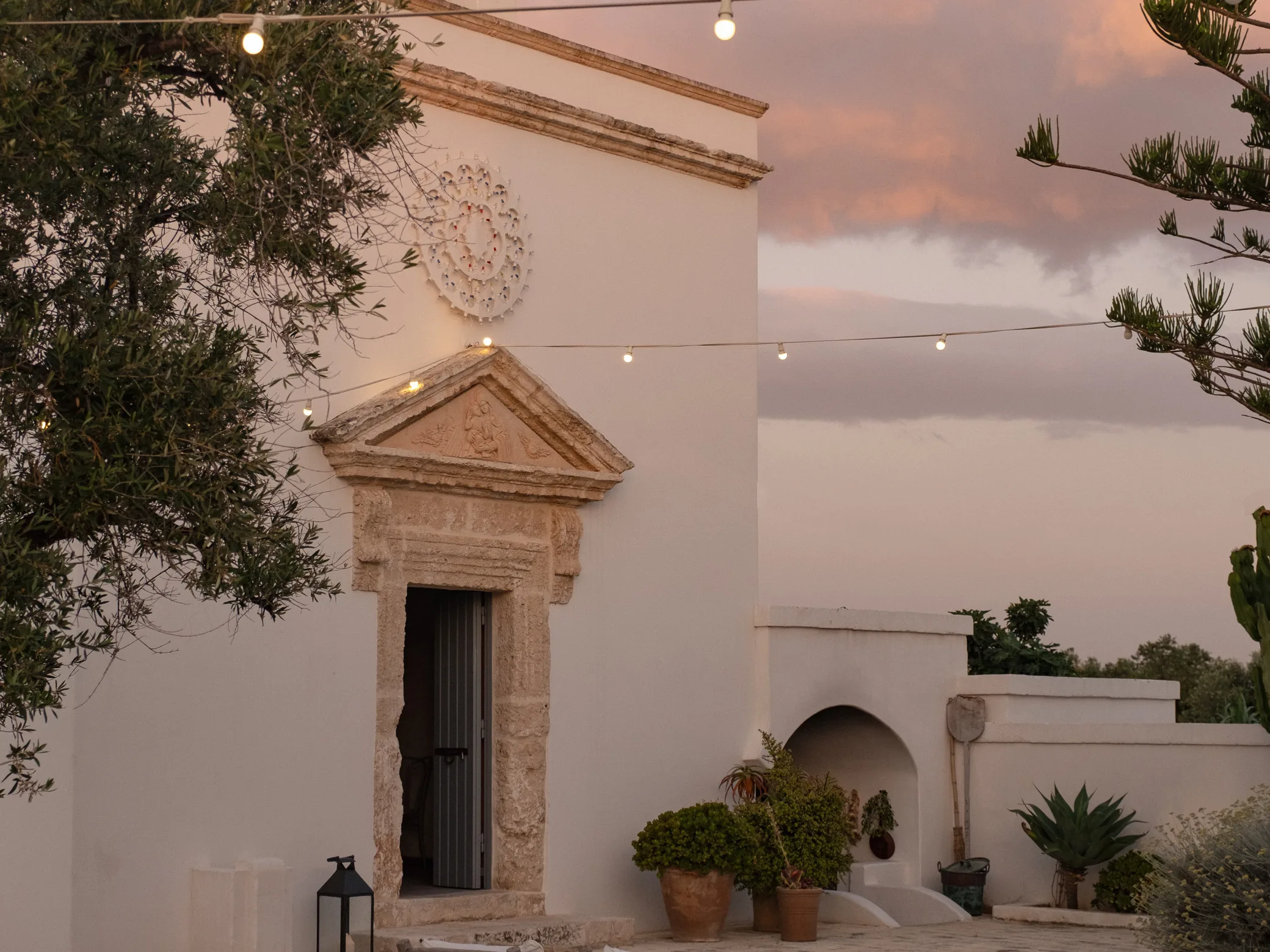 The chapel facade and lit courtyard at Masseria Calderisi at dusk, festoon lights strung above an empty terrace