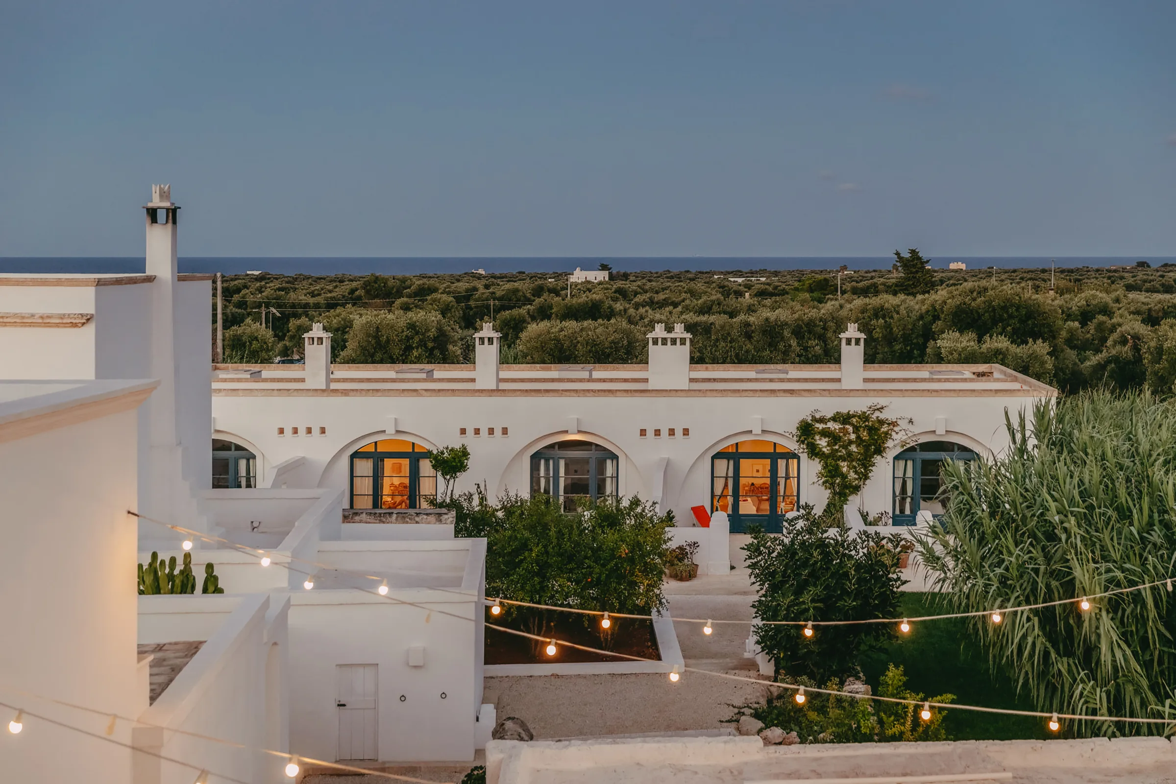 Masseria Calderisi at dusk, festoon lights above the arched facade with the olive groves and sea beyond