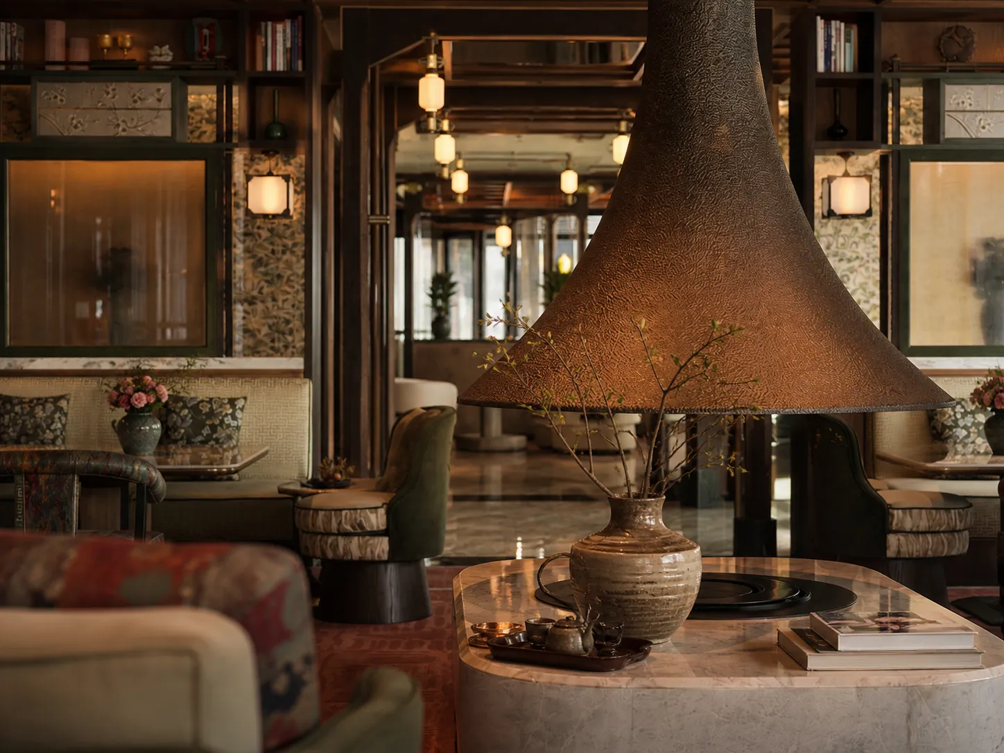 Layered Yunnan-modern hospitality interior — bronze pendant centrepiece, lantern lighting, patterned banquettes and a hand-thrown ceramic vessel